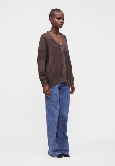 Femme debout portant un cardigan en maille marron ample avec des boutons et un jean bleu à jambes larges sur un fond clair uni.