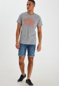 Camiseta gris con texto gráfico naranja, combinada con pantalones cortos de mezclilla azul. El modelo lleva zapatillas negras con suelas blancas, mostrando un atuendo casual.