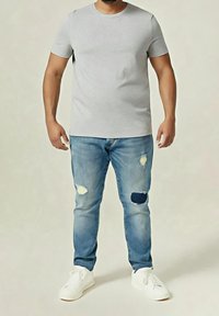 Homme portant un t-shirt gris clair à manches courtes, un jean bleu délavé avec des patchs et des baskets blanches, debout devant un fond uni.