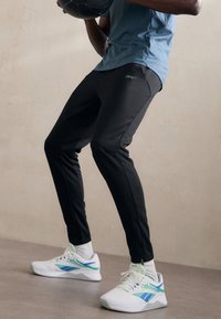 Sorte treningsbukser med slim fit; kombinert med hvite sko med grønne og blå detaljer. Stoffsammensetningen har en glatt overflate.