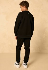 Schwarzer Sweater mit lässigem Schnitt, gerippten Bündchen und Logodetail auf dem Rücken. Kombiniert mit schwarzen Jogginghosen und weißen Sneakers. Glatte Stofftextur.