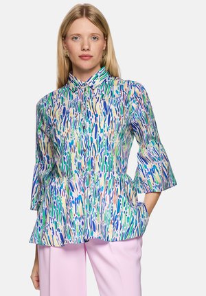 Blonde Frau trägt eine bunte Bluse mit abstraktem Muster, 3/4 Glockenärmel und helle rosa Hose, steht mit einer Hand in der Tasche.