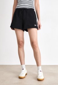 Sorte treningshorts med elastisk midje og logo-detalj. Kombinert med hvite joggesko og grå ankelstrømper, iført en klassisk stripete topp.