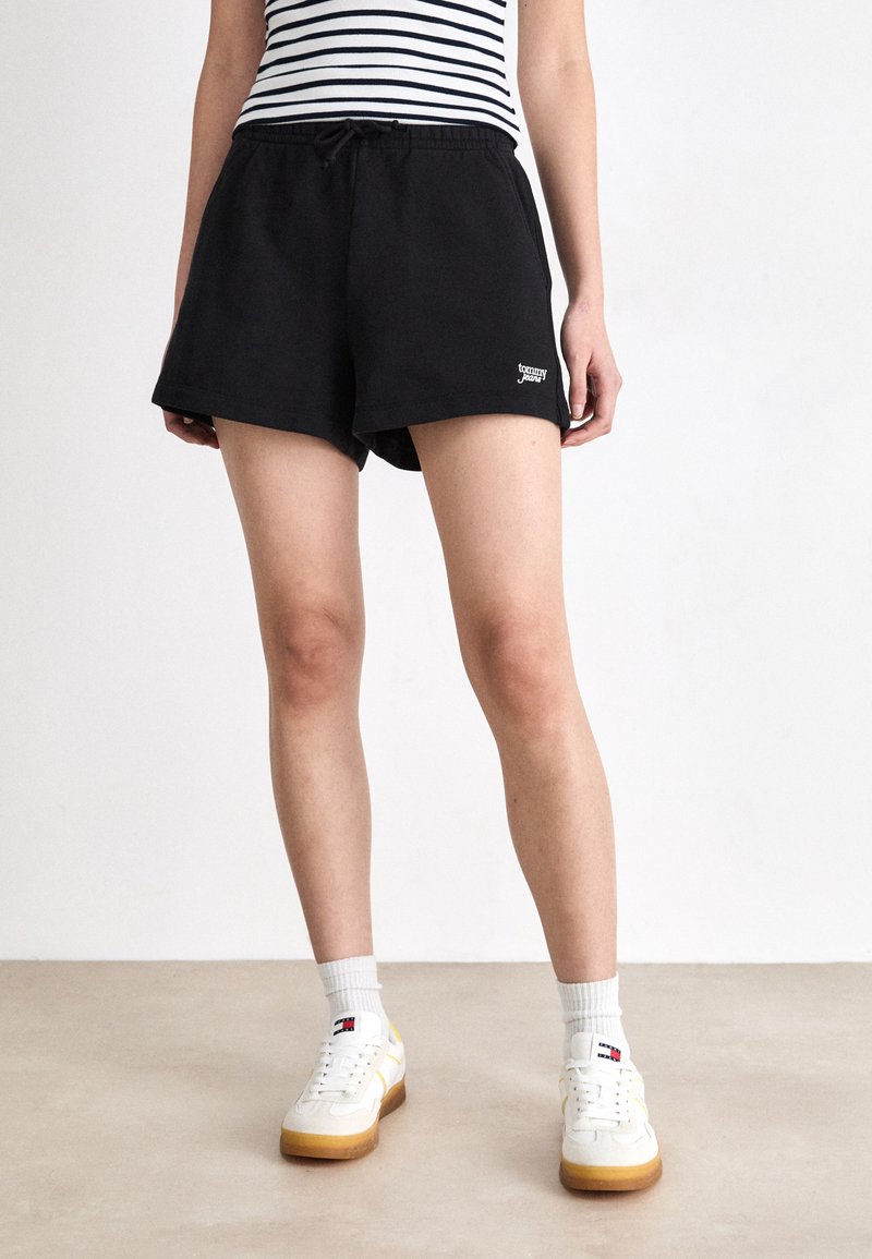 Sorte treningshorts med elastisk midje og logo-detalj. Kombinert med hvite joggesko og grå ankelstrømper, iført en klassisk stripete topp.