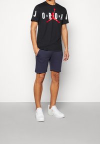 Czarna koszulka z krótkim rękawem z czerwoną i białą grafiką "JORDAN"; zestawiona z granatowymi szortami i białymi sneakersami. Prosty fason, materiał bawełniany.