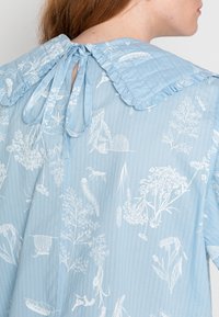 Blusa azul clara com colarinho oversized em babados e laço nas costas. Apresenta ilustrações botânicas brancas sobre um tecido texturizado.