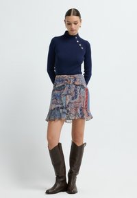 Marineblauwe geribbelde trui met een hoge nek en knoopdetail, gedragen met een kleurrijk gestreepte rok. Bruine leren laarzen tot aan de knie maken de look af.