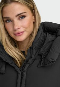 Sort dunjakke med høj krave og lynlås. Materialet er glat og skinnende med en puffet struktur omkring skuldrene.