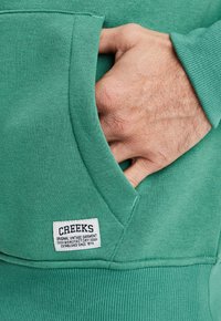 Sudadera verde con bolsillo frontal, puños acanalados y una etiqueta que dice "CREEKS" junto con detalles adicionales. Textura de algodón suave.