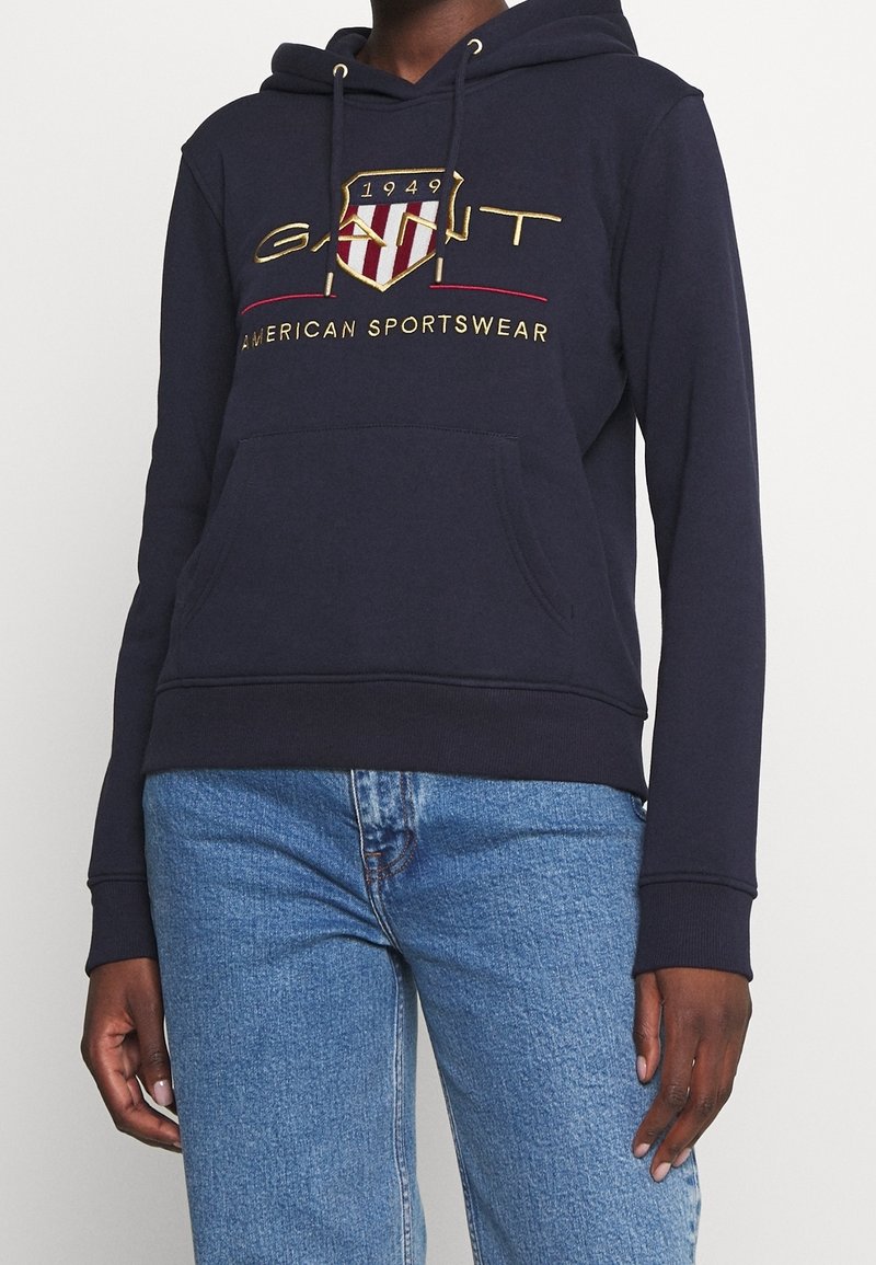 Personne portant un sweat à capuche bleu marine avec le logo "GANT 1949 American Sportswear" et un jean bleu clair, debout devant un fond uni.