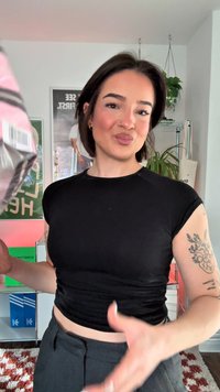 Schwarzes eng anliegendes Oberteil mit kurzen Ärmeln, kombiniert mit hoch taillierter, dunkler Hose. Im Hintergrund sind bunte Kisten und eine Zimmerpflanze zu sehen.