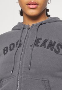 Haut à capuche zippé gris en texture polaire, avec le logo brodé "BDG JEANS" en noir, capuche avec cordon de serrage et poches avant. Tirette en métal argenté.