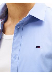 Hellblaues Hemd mit Knöpfen, Kragen und kurzen Ärmeln. Auf der Brust befindet sich ein gesticktes Logo mit roten, weißen und marineblauen Streifen.