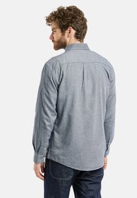 Homme aux cheveux bouclés et à la barbe, portant une chemise à manches longues gris clair et un jean foncé, vu de dos.