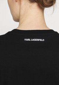 Svart bomullströja med ribbad rundhals och en vit "KARL LAGERFELD" logotyp tryckt på baksidan nära nacken.