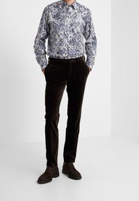 Mężczyzna w niebieskiej i białej koszuli z długim rękawem w kwiatowy wzór, w ciemnych brązowych aksamitnych spodniach i ciemnych brązowych butach na sznurówki, stoi z rękami w kieszeniach.