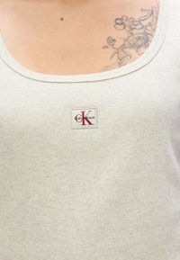 Ανοιχτό γκρι ριμπ αμάνικο τοπ με φαρδύ λαιμόκοψη και τετράγωνο λογότυπο "Calvin Klein" σε κόκκινο και μαύρο.