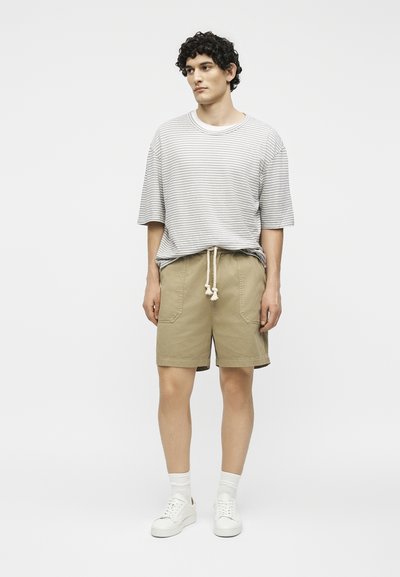 Νέος άνδρας στέκεται φορώντας μια γκρι ριγέ μπλούζα, μπεζ σορτς με ζώνη σχοινί, λευκές κάλτσες και λευκά αθλητικά παπούτσια σε απλό ανοιχτό φόντο.