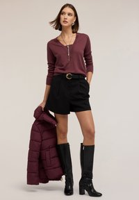 Maroon v-paita, mustat korkeavyötäröiset shortsit vyöllä, polvipituiset mustat saappaat, ja marooni toppatakki pidettään yhdessä kädessä.