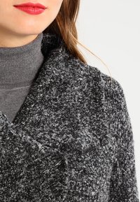 Grauer, strukturierter Mantel mit weitem Kragen, aus weichem Stoff, mit einem gesprenkelten Muster. Darüber ein dunkler Rollkragenpullover getragen.