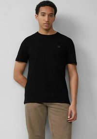 Schwarzes Baumwoll-T-Shirt mit kurzen Ärmeln, rundem Ausschnitt und einem dezenten Logodetail auf der Brust. Kombiniert mit hellen Hosen.