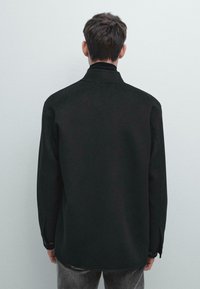 Maglietta nera realizzata in un tessuto testurizzato, con un design semplice che presenta un colletto e polsini con bottone. La vestibilità è ampia e leggermente oversize.