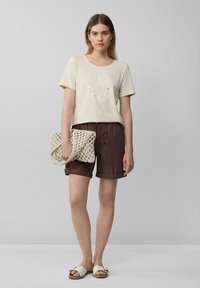 T-shirt beige à manches courtes avec design graphique, shorts marron à ourlet retroussé, sac à main en tissé crème, et sandales blanches.