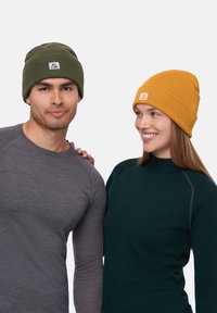 Gorros de punto en verde y mostaza con un acabado texturizado. Etiquetas con un logo. El modelo lleva camisetas de manga larga ajustadas en gris y verde.