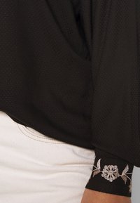 Top nero testurizzato con maniche lunghe, caratterizzato da ricami floreali metallici sui polsini. Indossato sopra un tessuto chiaro.