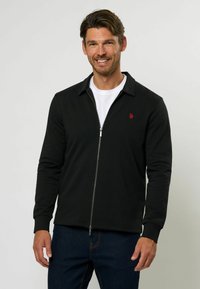 Melnas zip-up jaka ar norādītu apkakli, ribotām piedurknēm un mazu sarkanu logotipu kreisajā krūtī, uzvilkta virs baltas krekla.