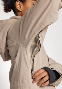 Chaqueta de color beige claro con detalle de cremallera y forro de malla, que presenta puños elásticos en color marino. Tejido suave y corte ajustado con capucha.