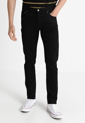 Man wearing slim-fit black jeans with white sneakers, one hand in pocket, against a plain white background.