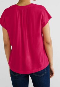Top corto fucsia de manga corta con cuello redondeado, detalle de pliegues en la parte trasera, textura suave y un corte relajado, combinado con jeans azul oscuro.