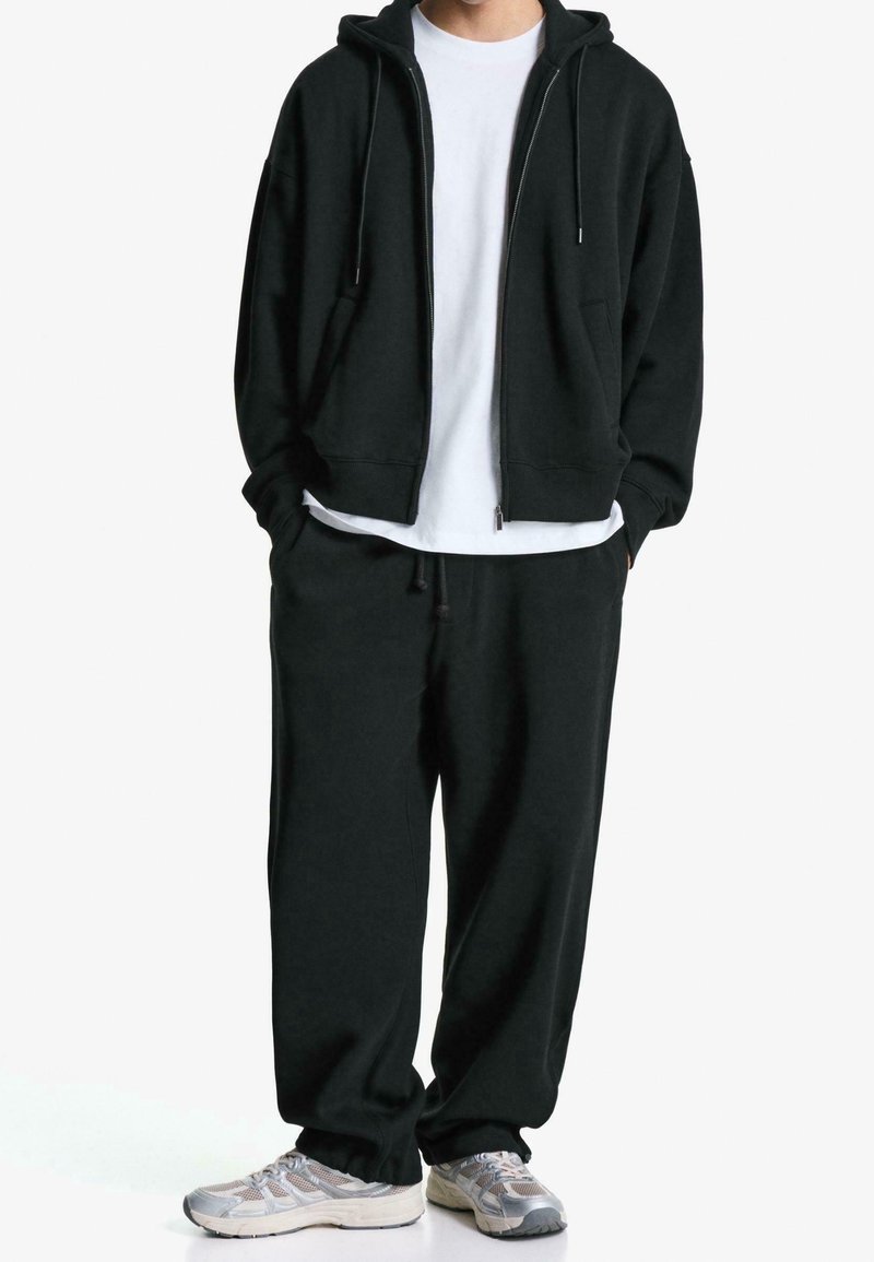 Personne portant un sweat à capuche noir zippé et un pantalon de survêtement noir assorti, avec des baskets blanches et un T-shirt blanc en dessous.