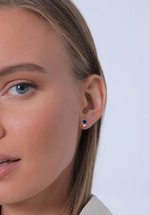 Tropfenohrringe mit einem blauen Edelstein, der von kleinen klaren Akzenten umgeben ist, in einer Metallfassung, die am Ohrläppchen getragen wird.