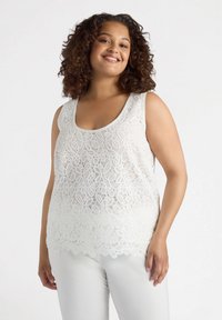 White lace tank top with a scoop neckline, featuring intricate floral patterns and a scalloped hem. Paired with white pants.
