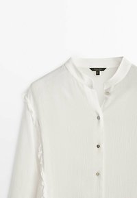 Blouse blanche texturée à manches longues avec petit col montant et trois boutons visibles sur le devant.