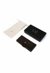 Donkerbruine suède portemonnee met een gesp, vergezeld van een zwarte doos en een crèmekleurig trekkoordzakje met het label "L37." Gladde textuur en minimalistisch ontwerp.