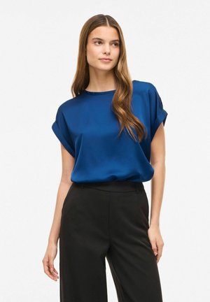 Femme portant un chemisier bleu en satin à manches courtes, rentré dans un pantalon noir taille haute, debout devant un fond blanc uni.