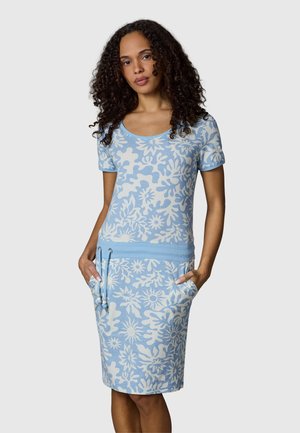 Kvinne med krøllete hår iført en lyseblå og hvit blomstrete kortermet kjole med lommer og snøring i midjen, står foran en grå bakgrunn.