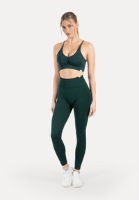 Donkergroene sport-bh met geplooide details en bijpassende hoge taille legging. Beide gemaakt van glad, rekbaar materiaal. Witte sneakers gedragen.