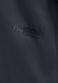 Ciemna tkanina z bliska z wytłoczonym logo "hummel PERFORMANCE" na fakturowanym, gładkim materiale sportowym.