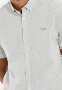 Camisa de manga corta de color gris claro, hecha de una tela texturizada, con un cierre de botones en la parte delantera y un pequeño logotipo bordado en azul marino en el lado izquierdo del pecho.