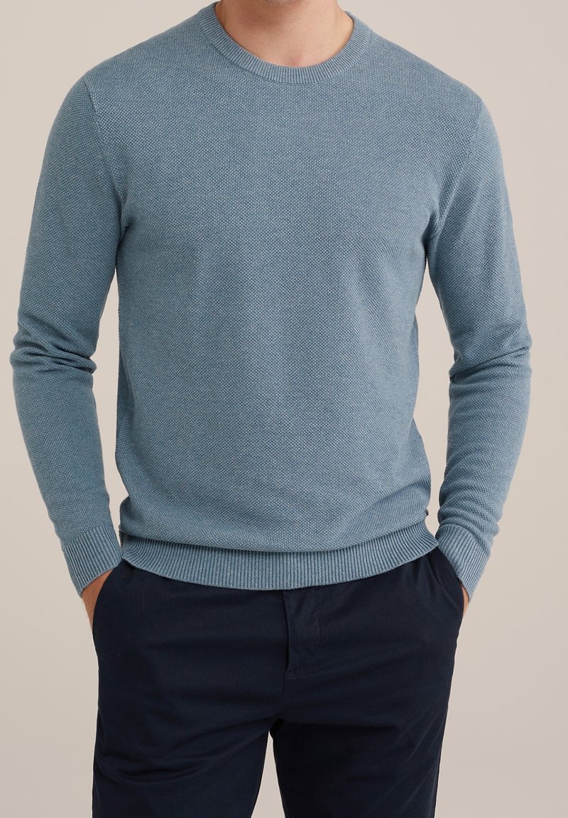 Man i en ljusblå strukturerad stickad tröja med långa ärmar och mörkblå byxor, stående med händerna i fickorna.