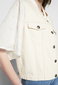 Colete de ganga bege cortado com bolsos frontais, botões prateados e uma manga branca com rendas. Apresenta um design estruturado e um tecido texturizado.