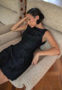 Musta ilmanhihoinen mekko, jossa on teksturoitu kangas, sovitettu siluetti ja risattu helma. Malli rentoutuu beessissä punottu tuolissa, jossa on puuakcentteja.