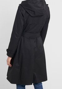 Svart vattentålig kappa med huva, bälte och knäppta ärmslut; har en slät, matt yta och en utsvängd form i fållen.