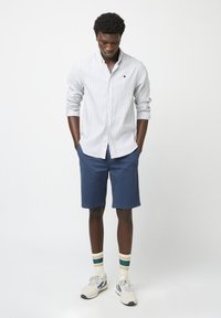 Camisa de hombre de botones en rayas azul claro y blanca, combinada con pantalones cortos azul oscuro. Lleva zapatillas blancas y calcetines a rayas.