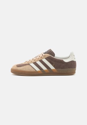 Hnědý a béžový teniska Adidas Gazelle s bílými pruhy, gumovou podrážkou a texturovaným hnědým jazykem, zobrazená z boku na bílém pozadí.