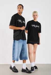 Svart grafisk t-skjorter med hvite design, denimshorts og svarte sko. En modell bærer en liten svart veske. Enkelt, uformelt gateklesstil.